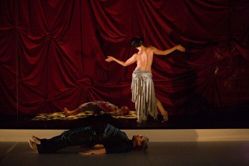 Two figures lying on the stage floor with one other standing with their bare back to the audience