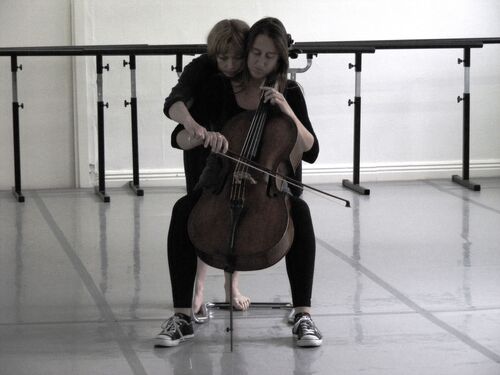 A figure playing cello while another leans around to hold her bowing hand