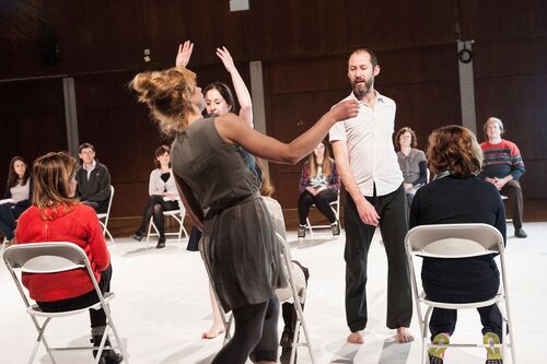 Three dancers in movement among audience seated to the side of the performance space