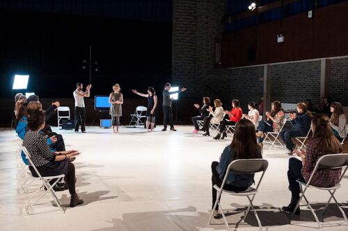 An ensemble of dancers standing with audience on 3 sides of the performance space