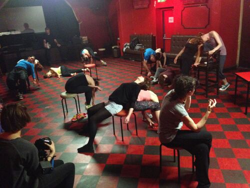 An ensemble of participants in movement over chairs scattered throughout a room