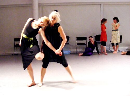 Two participants in movement together in a studio space with three others standing in the background