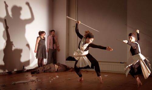 An ensemble of participants in movement, two with long staffs in hand casting shadows behind them