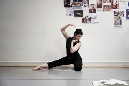 A figure in movement in a studio with images stuck to a plain white wall behind