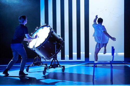 One figure at a large drum and another figure in movement on a blue lit stage