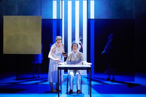 A figure seated at a table with another figure reaching for her hand centre stage