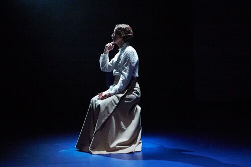 A figure in a long skirt seated in side profile on stage