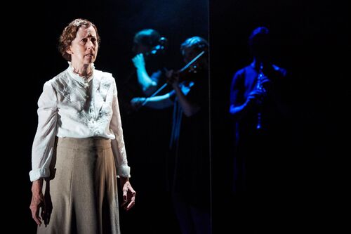 A figure standing on a dark stage with figures lit in blue in the background