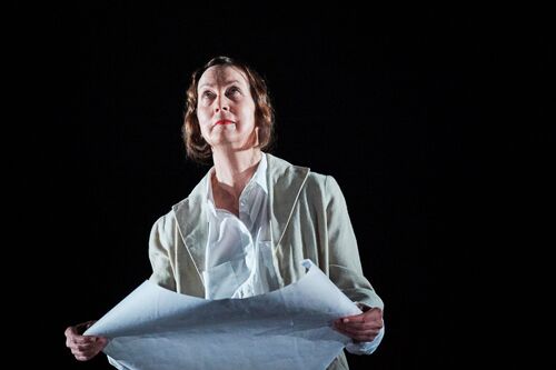 A figure dressed in white holding a roll of paper and looking upwards on a dark stage