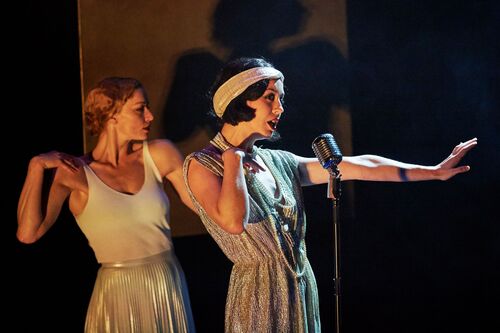 Two figures, one standing at a microphone and the other mirroring her movement behind