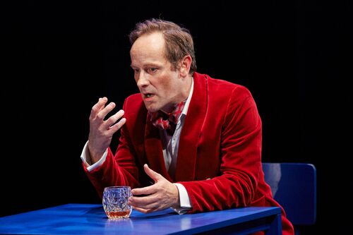 A figure in red seated at a blue table on stage