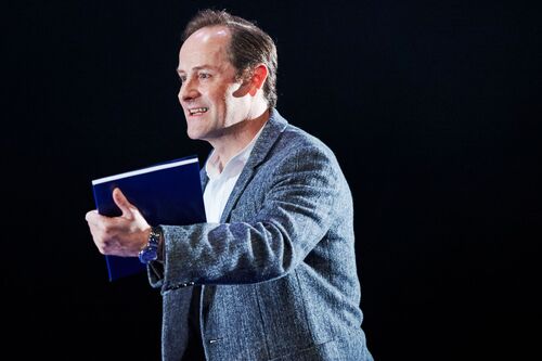 A figure holding a blue book in hand on a dark stage