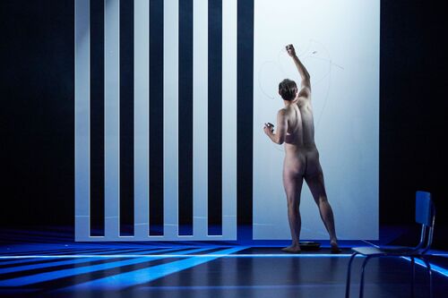 A nude figure with their back to the audience in movement on stage