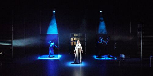 A figure standing spotlit on a dark blue stage with other figures in movement in the background