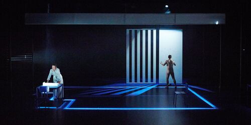 Two figures on a dark stage lit with blue accents, one facing away from the audience