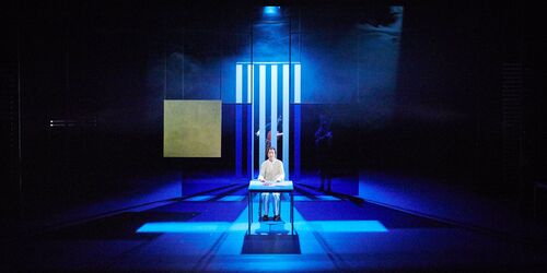 A seated figure in white in the centre of a blue lit stage