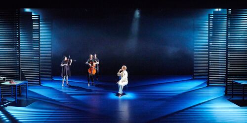 Three musicians and a seated actor in the centre of a stage lit in blue