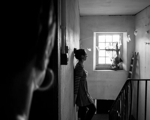 A figure in side profile at the top of a flight of stairs seen over the shoulder of another person