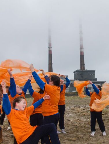 A group of children in orange tshirts and rain ponchos in movement in front of the Poolbeg Chimneys