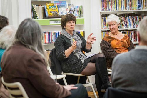 A person speaking to a group of people seated in a circle with bookshelves surrounding