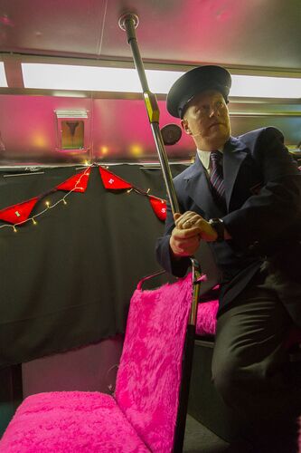 A figure costumed as a conductor on the upper deck of a bus with bright pink seats