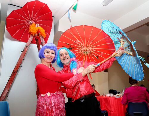 A couple in brightly coloured wigs holding red and blue parasols and smiling to camera