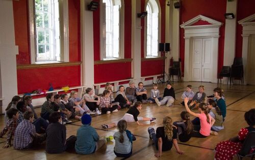 A circle of children seated with one child doing the splits in the centre of the circle