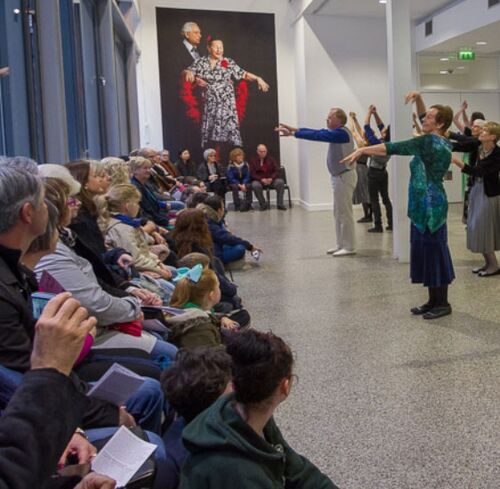 Participants seen from the side performing for an audience seated in a gallery space