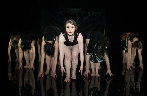 An ensemble of participants, all bent over with heads down except for one figure facing front