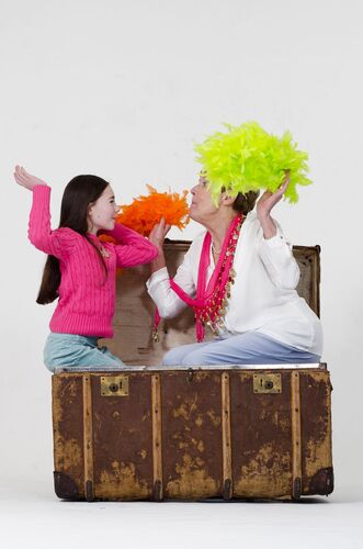 An older figure and a child sitting in a treasure chest with feather boas facing each other