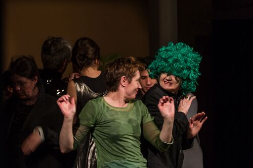 Participants in movement in a tight huddle, one with a bright green curly wig on her head