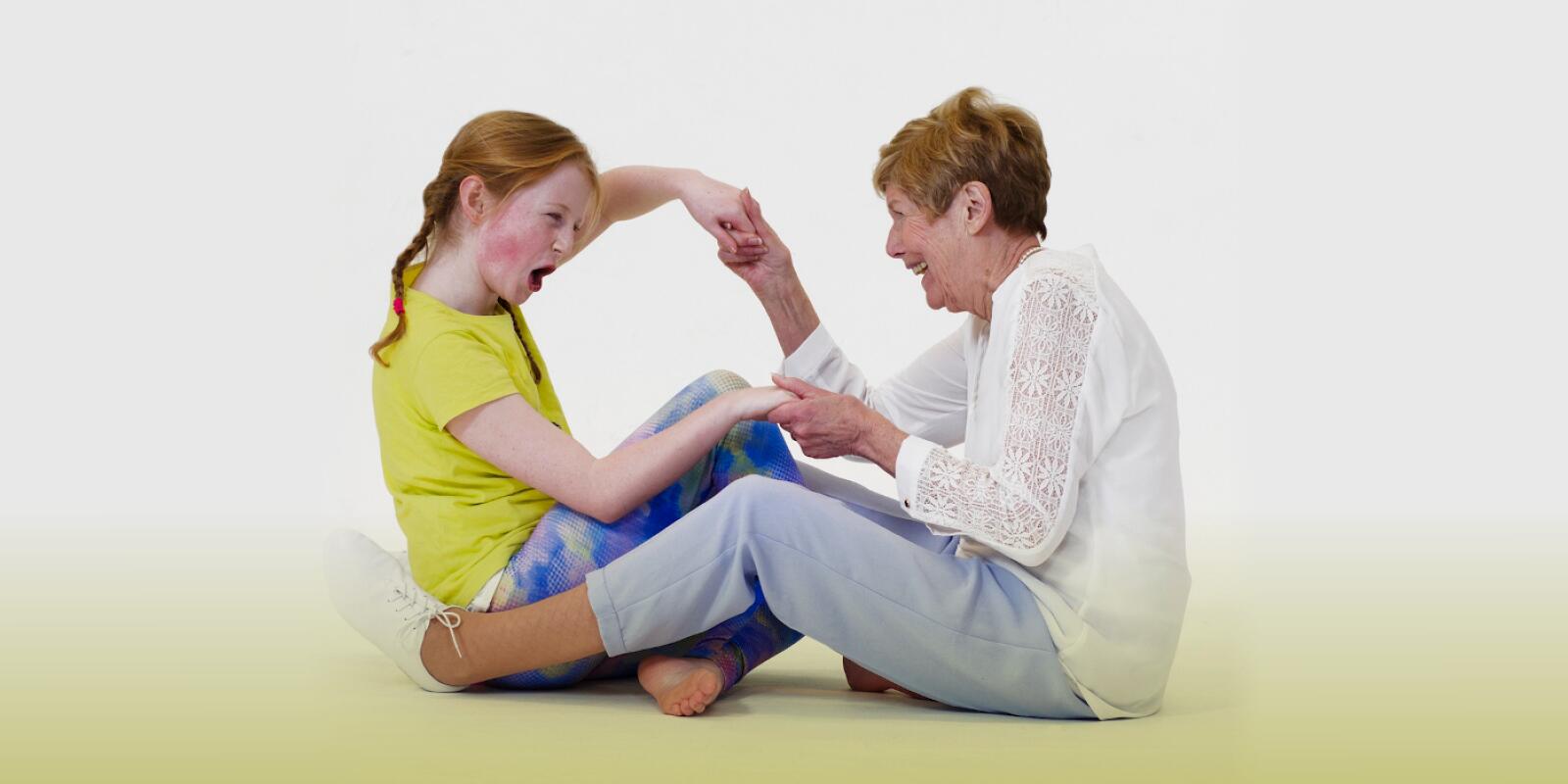 An older figure and a child sit opposite each other holding hands and laughing