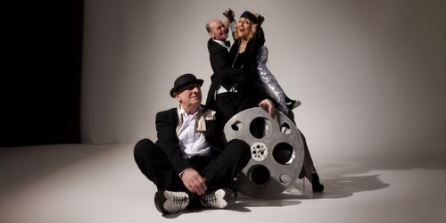 Two figures dancing with another seated cross-legged at their feet beside a huge film reel