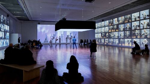 An ensemble of dancers moving in a gallery space surrounded by audience throughout the room
