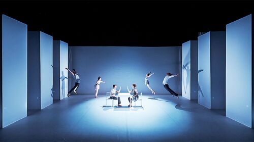 Two figures seated opposite each other centre stage with four other figures in movement around them