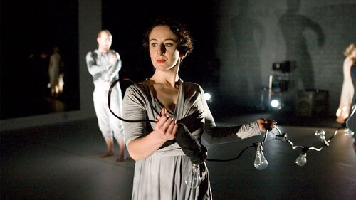 A figure standing holding a string of festoon lightbulbs behind