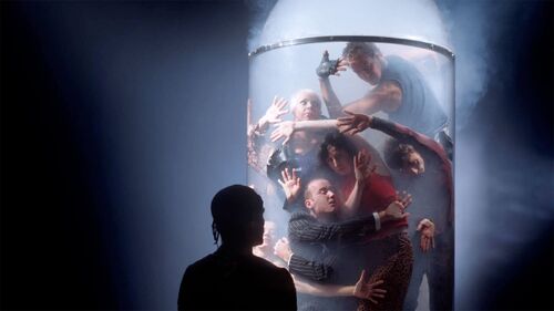 An ensemble of dancers pressed against the insdie of a glass cylinder full of haze