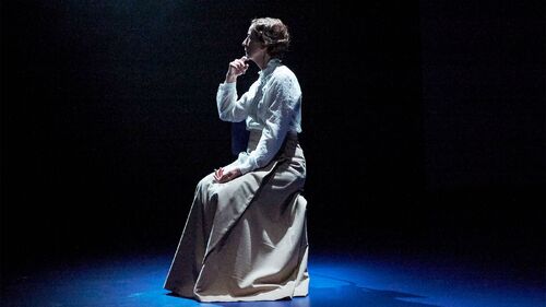 A figure in a long skirt sitting in side profile centre stage