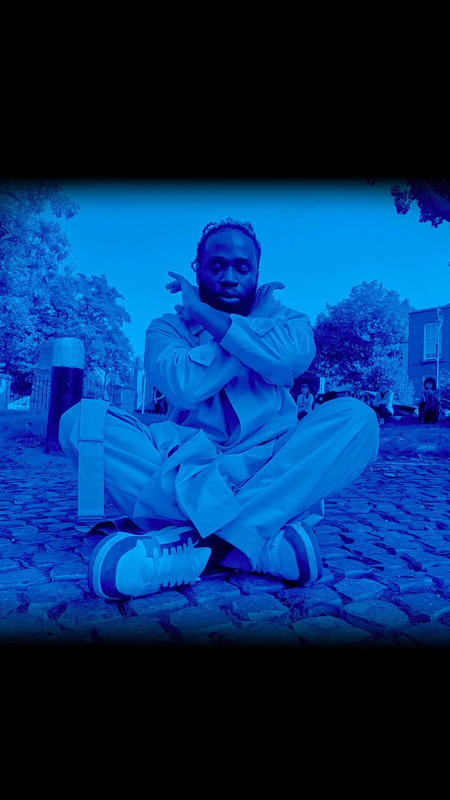 A blue filtered image of the dancer sitting on the ground in a pose with crossed legs and arms