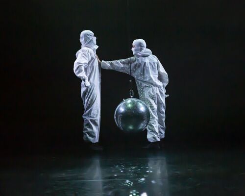 A couple of dancers in white hazmat suits and balaclavas interacting around a mirror ball